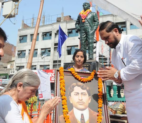 A garlanded framed portrait of Madan Lal Dhingra set before his memorial statue during a public tribute, with flags and marigold garlands and people standing around it.