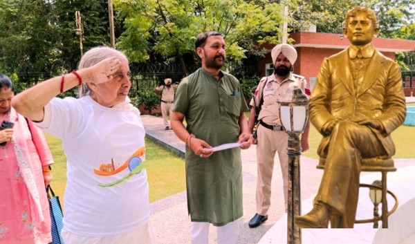 A gilded statue of Madan Lal Dhingra seated in a chair in a memorial park, with visitors and a uniformed police officer standing nearby.