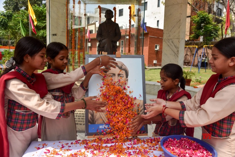 Four schoolgirls in uniform scatter marigold and rose petals over a framed portrait of Madan Lal Dhingra at an outdoor memorial, with a bronze seated statue in a glass pavilion behind.
