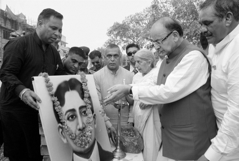 Black and white photograph of a commemorative ceremony in which a tall brass lamp is lit before a garlanded framed portrait of Madan Lal Dhingra.