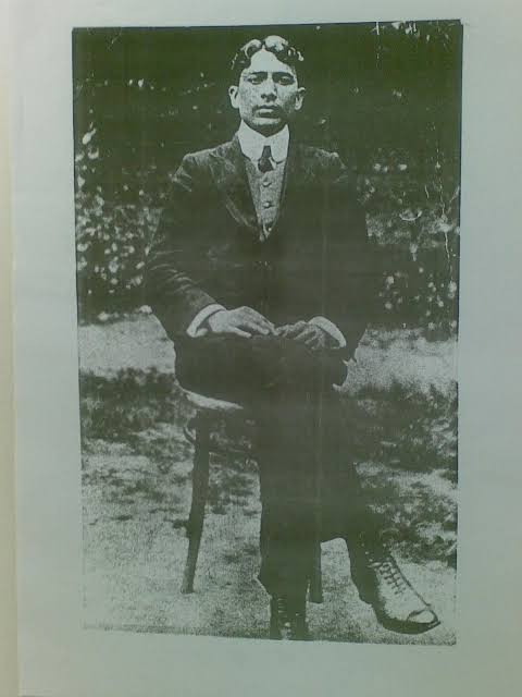 Full-length photograph of Madan Lal Dhingra seated on a bentwood chair in a garden, wearing a dark three-piece suit and boots.